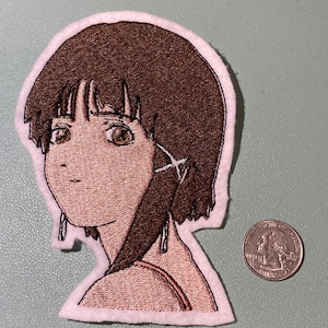 Può includere: Toppa ricamata raffigurante un personaggio in stile anime con capelli e occhi marroni, che indossa un top marrone chiaro. La toppa ha un bordo bianco ed è accanto a una moneta come riferimento alle dimensioni. La toppa è su una superficie verde chiaro.