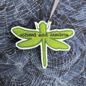 May include: A lime green embroidered dragonfly patch with a white border and the text coheed and cambria stitched in black across the wings. The patch is held by a pair of silver metal tweezers against a dark blue spider web fabric.