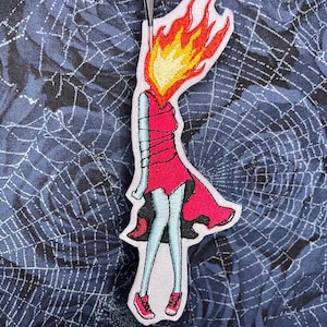 May include: An embroidered patch featuring a figure with a flame for a head, wearing a bright pink dress and light blue limbs. The figure is wearing pink sneakers and is held by a pair of metal tweezers, showcasing detailed stitching and vibrant colors.