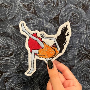 May include: Embroidered patch featuring a stylized figure in a red dress and orange skirt, with black hair. The patch has a white border and is set against a dark blue fabric with a spiderweb pattern. The figure is in a dynamic pose.