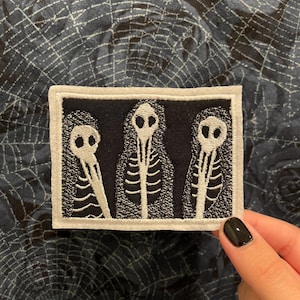 May include: A rectangular embroidered patch featuring a black background with three white skeletal figures. Each figure has a bird-like skull head and a visible rib cage design. The patch is framed by a thick white border with stitched edges.