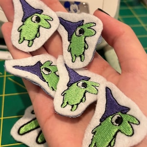 May include: A collection of embroidered patches featuring a green creature with a purple witch hat. The patches are on a white background with a stitched border. The creature has a large eye and is standing upright.