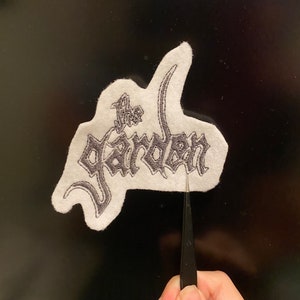May include: White felt patch with black embroidered text that reads "The Garden".