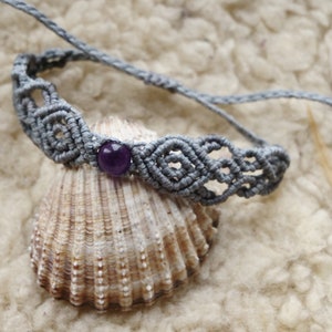 May include: A gray macrame bracelet with a purple amethyst bead. The bracelet is on a seashell.