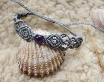 Puede incluir: Un brazalete de macramé gris con una cuenta de amatista púrpura. El brazalete está sobre una concha.