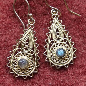 May include: A pair of gold dangle earrings with a teardrop shape. Each earring features a delicate filigree design and a round, iridescent gemstone in the center.