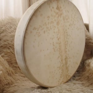 May include: A large, round drum with a light-colored, spotted surface. The drum has a wooden frame and is resting on a fluffy, cream-colored surface. The background features a soft, neutral-toned curtain.
