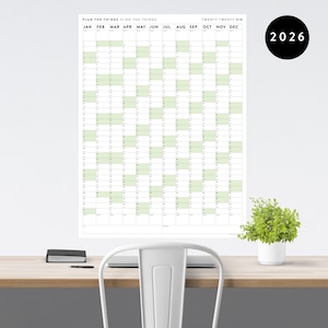 May include: A large wall calendar for the year 2026, with a white background and a grid layout. The calendar features green-colored boxes for each day and the text "PLAN THE THINGS // DO THE THINGS". A small plant in a white pot sits on a wooden desk.