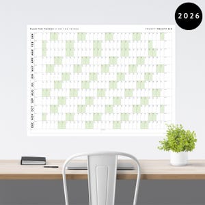 Peut inclure: Un grand planificateur annuel blanc de 2026 avec des dates en vert est affiché sur un mur blanc. Le planificateur porte l'inscription "PLAN THE THINGS // DO THE THINGS" et "TWENTY TWENTY SIX". Une petite plante en pot blanc est posée sur un bureau en bois.