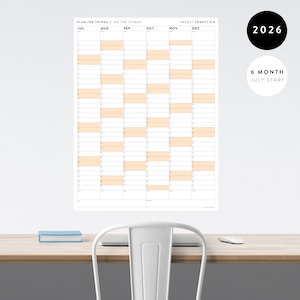 May include: A large white wall calendar for 2026, with a 6-month July start. The calendar features a grid layout with monthly columns and rows for each day. The text "PLAN THE THINGS // DO THE THINGS" is at the top. The calendar is hanging on a white wall.
