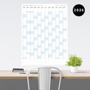 Peut inclure: Un grand calendrier mural blanc pour l'année 2026, avec un design minimaliste. Le calendrier présente une mise en page en grille avec des cases bleues claires en surbrillance pour chaque mois. Le texte "PLAN THE THINGS // DO THE THINGS" est en haut.
