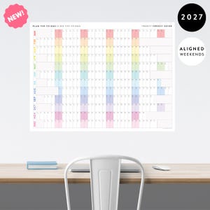 May include: A 2027 wall calendar featuring a horizontal grid layout with months listed vertically in rainbow colors. The header reads PLAN THE THINGS // DO THE THINGS and TWENTY TWENTY SEVEN. It includes a pink NEW badge and a white ALIGNED WEEKENDS circle.