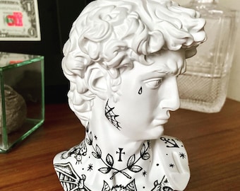 May include: White plaster bust of a man with black and white tattoos on the neck and chest. The tattoos include a cross, bats, and an anchor.