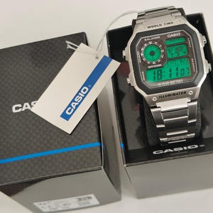 May include: A silver Casio digital watch with a black face and green digital display, resting on a black box. The watch features the text "WORLD TIME" and "ILLUMINATOR". A white tag with the Casio logo is attached to the box.