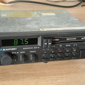 Puede incluir: Una radio de coche Blaupunkt Woodstock SQR 06 vintage, con una pantalla digital que muestra "87.5". La placa frontal negra tiene botones para bajos, agudos y presintonías. Se ve un reproductor de casetes. La radio está sobre una superficie de madera.