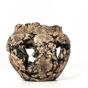 Op de afbeelding: Een bruine keramische plantenpot met een verweerde afwerking en verschillende uitgesneden gaten. De pot heeft een ronde vorm en een ruwe textuur.