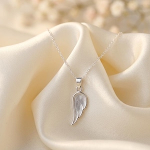 May include: A silver-colored pendant necklace featuring a wing-shaped charm. The wing has a textured surface and hangs from a delicate chain. The necklace is displayed on a cream-colored satin fabric.