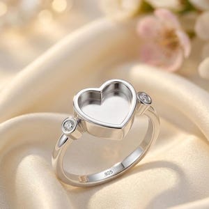 Könnte beinhalten: Ein Ring aus Sterlingsilber mit einer herzförmigen Fassung in der Mitte, die für einen Stein oder eine Einlage vorgesehen ist. Der Ring ist mit .925 gestempelt und hat zwei kleine, runde, klare Schmucksteine an den Seiten des Herzens.