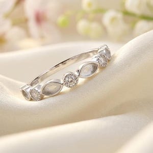 May include: A silver ring featuring alternating round and marquise-shaped settings, each set with small, sparkling stones. The ring is displayed on a soft, cream-coloured fabric, with blurred floral elements in the background.