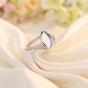 Könnte beinhalten: Ein silberfarbener Ring mit einer zentralen, marquiseförmigen, leeren Fassung für einen Edelstein. Die geteilte Ringschiene ist mit kleinen, klaren, im Pavé-Stil gefassten Steinen besetzt, die sich entlang beider Seiten des Rings erstrecken.