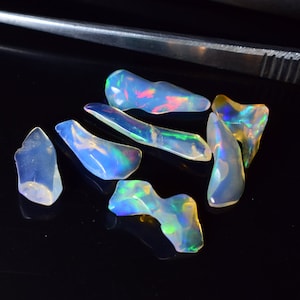 May include: A group of seven iridescent opal gemstones in various shapes and sizes. The opals are a mix of white, blue, green, and yellow.