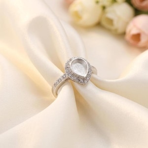 Op de afbeelding: Een zilverkleurige ring met een peervormige zetting ontworpen voor een edelsteen. De ringband en de halo rondom de centrale peervormige cup zijn ingelegd met kleine, heldere, fonkelende steentjes die het licht opvangen en weerkaatsen.