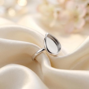 Könnte beinhalten: Ein silberner Ring mit tropfenförmiger Fassung, auf cremefarbenem Satin-Stoff. Die Fassung des Rings ist leer, bereit für einen Stein oder eine andere Verzierung. Weichzeichner-Hintergrund mit floralen Elementen.
