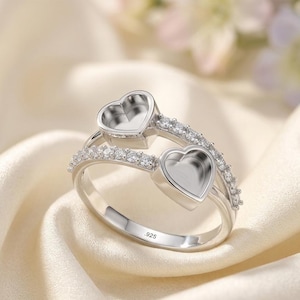 Könnte beinhalten: Ein silberfarbener Ring mit zwei leeren herzförmigen Fassungen und einem Band, das mit kleinen klaren Steinen besetzt ist. Die Innenseite des Rings ist mit dem Stempel .925 versehen, was auf eine Zusammensetzung aus Sterlingsilber hinweist.