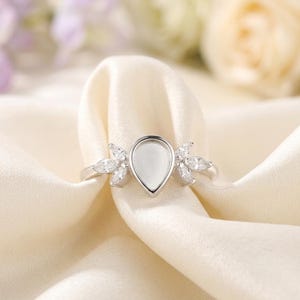 Peut inclure: Une bague en argent avec un sertissage en forme de poire, entourée de petites pierres précieuses transparentes. La bague repose sur un tissu crème, avec des fleurs floues en arrière-plan. Le design de la bague est élégant et délicat.