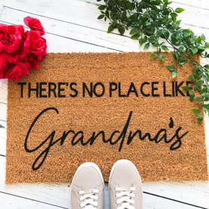 Puede incluir: Una alfombra de coco marr&oacute;n con el texto "There's no place like Grandma's" en letras negras. La alfombra est&aacute; sobre una superficie de madera blanca.