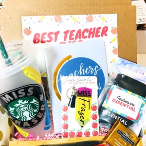 May include: A gift box filled with teacher appreciation gifts, including a reusable cup with a green logo, a notebook with the words "Teachers you hold the hope of growth", a keychain with a pencil charm, a chocolate bar, and a red pen.
