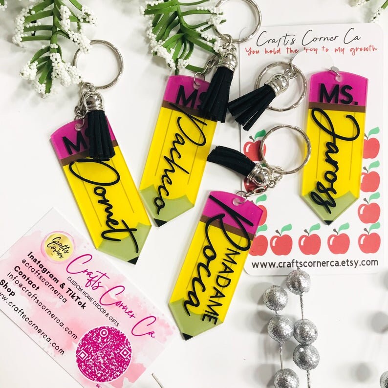 May include: Four yellow and pink pencil-shaped keychains with black tassels. Each keychain has a different name on it: "Ms. Domet", "Ms. Pacheco", "Ms. Jarosz", and "Madame Rocca". The keychains are on a white surface with a business card that says "Crafts Corner Ca" and a website address.