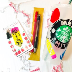 May include: A teacher appreciation gift set with a personalized pencil keychain, two pens, and a Starbucks cup with a teacher-themed design. The cup has a green Starbucks logo with the text "MRS. SMITH" on it. The pencil keychain is yellow with the text "MRS. SMITH" on it.