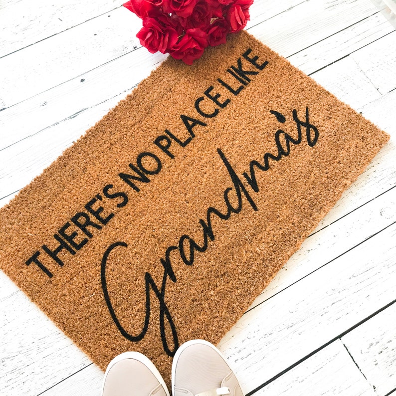 Puede incluir: Una alfombra de coco marr&oacute;n con el texto "There's no place like Grandma's" en letras negras.