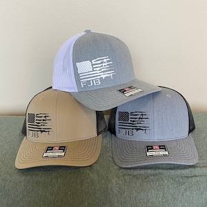 May include: Three baseball caps with a trucker hat style are displayed. Each hat features a flag design with the letters "FJB" and a gun graphic. The hats are in gray, tan, and gray with white, black, and black mesh backs.