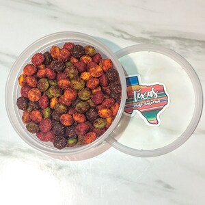 May include: A clear plastic container filled with colorful, seasoned candies. The candies are a mix of red, green, and brown, with a textured coating. The lid is partially open, revealing a sticker with the text "Texas chilidulceria" in a Texas-shaped design.