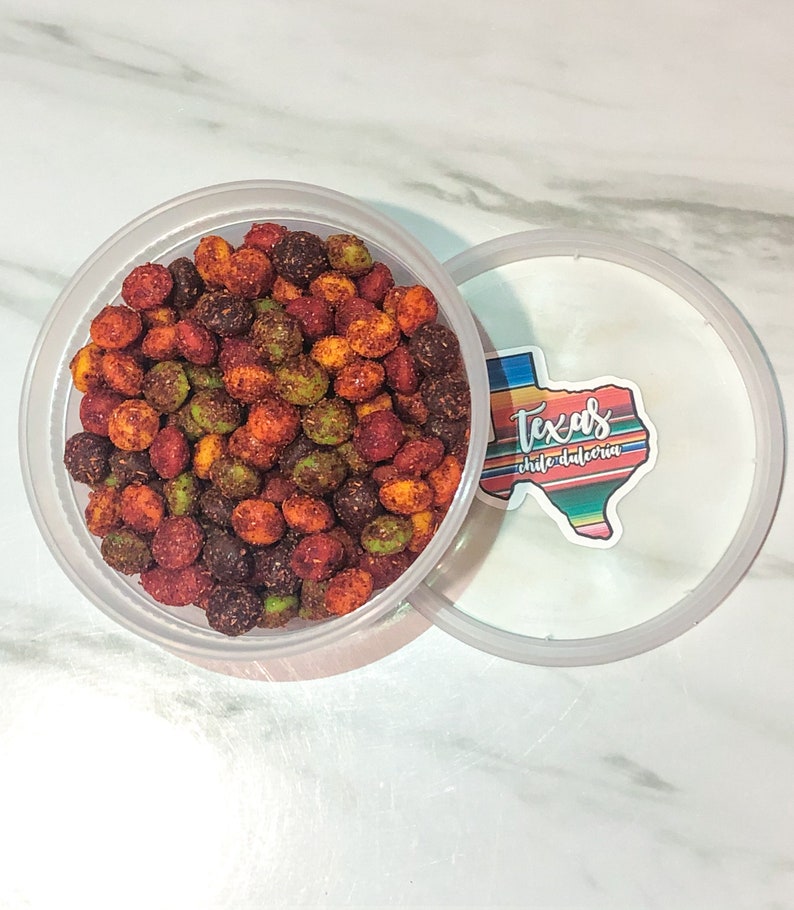 May include: A clear plastic container filled with colorful, seasoned candies. The candies are a mix of red, green, and brown, with a textured coating. The lid is partially open, revealing a sticker with the text "Texas chilidulceria" in a Texas-shaped design.