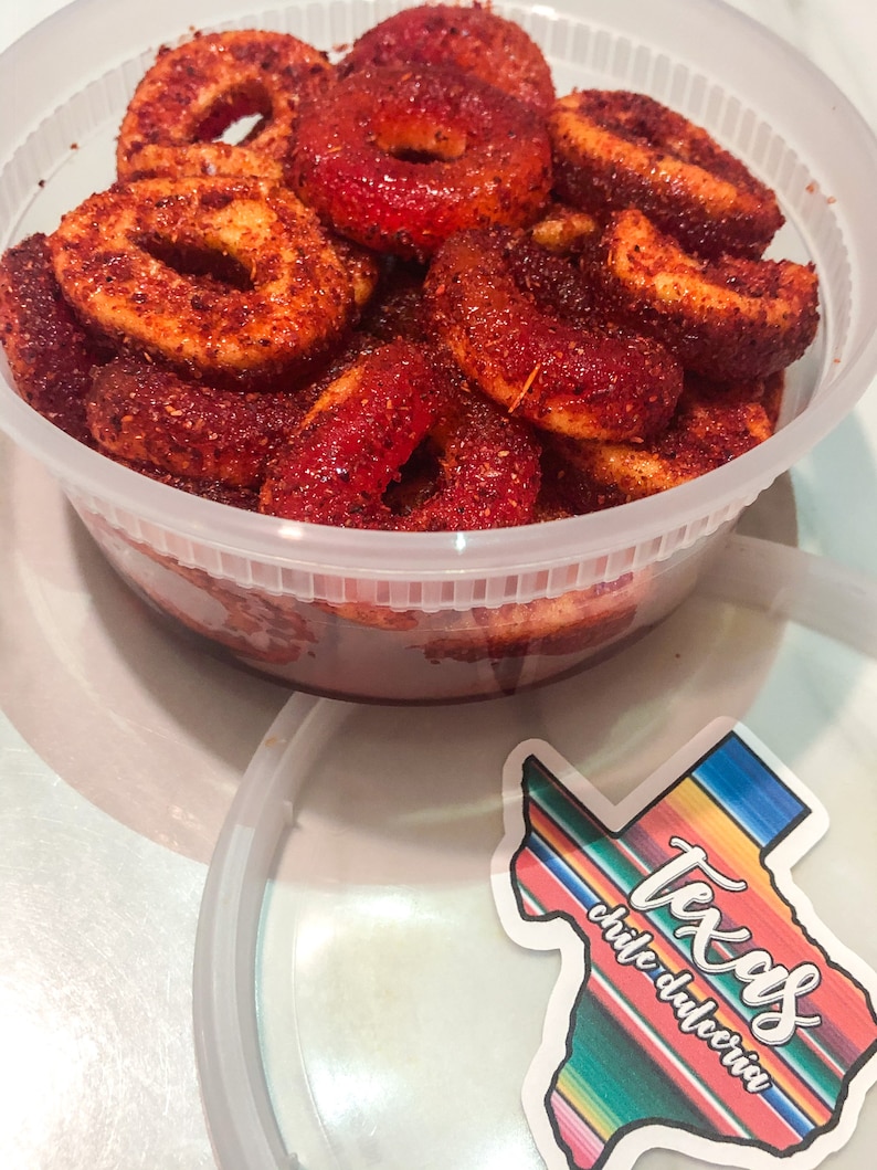 Puede incluir: Un recipiente de pl&aacute;stico transparente lleno de anillos de caramelo rojos y naranjas, recubiertos de chile en polvo. Una pegatina con el texto "Texas chile dulceria" en un dise&ntilde;o colorido est&aacute; al lado del recipiente.