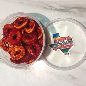 Puede incluir: Un recipiente de pl&aacute;stico transparente lleno de caramelos en forma de anillo espolvoreados de rojo. La tapa, parcialmente abierta, muestra una pegatina con el contorno de Texas y las palabras "Texas chile dulceria" en un dise&ntilde;o colorido. Los caramelos parecen ser un dulce y picante.