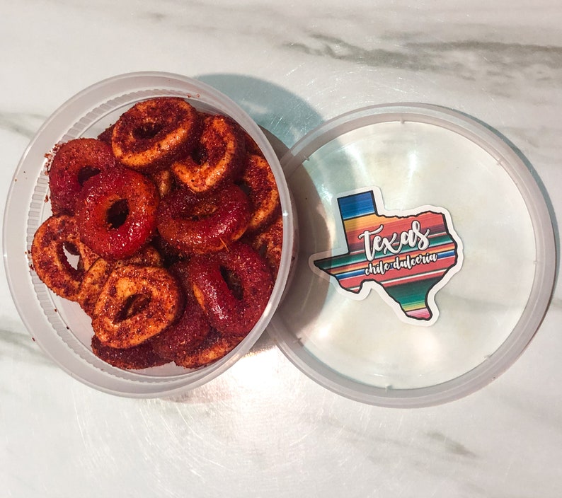 Puede incluir: Un recipiente de pl&aacute;stico transparente lleno de caramelos en forma de anillo espolvoreados de rojo. La tapa, parcialmente abierta, muestra una pegatina con el contorno de Texas y las palabras "Texas chile dulceria" en un dise&ntilde;o colorido. Los caramelos parecen ser un dulce y picante.