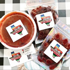 Puede incluir: Un surtido de dulces y salsas mexicanos. Un recipiente blanco de salsa roja est&aacute; junto a dos bolsas transparentes de dulces y una tarjeta de visita. Cada art&iacute;culo tiene una pegatina con el texto "Texas chile dulceria" en forma de Texas.