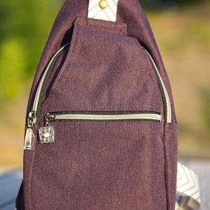 May include: Plum-colored sling backpack with a front zippered pocket and a decorative zipper pull. The bag has a white and gold accent on the top and a patterned strap. The background is blurred, suggesting an outdoor setting.