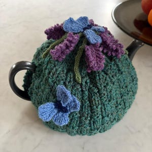 Könnte beinhalten: Eine teal-farbene Strick-Teekanne mit blauen und lila Blumenverzierungen. Die Kanne hat ein strukturiertes Strickmuster und ist mit gehäkelten Blumen und Blättern verziert.