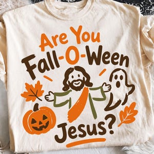 May include: A cream-colored t-shirt featuring a graphic print with the text Are You Fall-O-Ween Jesus? The design includes a central figure of Jesus, a ghost, a jack-o'-lantern pumpkin, and two autumn leaves in shades of orange and brown.