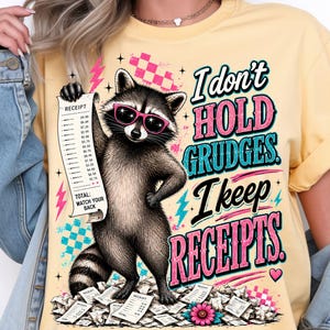 K&ouml;nnte beinhalten: Ein hellgelbes T-Shirt mit einer Grafik eines Waschb&auml;ren, der eine rosa Sonnenbrille tr&auml;gt und einen langen Kassenbon h&auml;lt. Der Text lautet, I don't hold grudges. I keep receipts. Das Design enth&auml;lt rosa und blaue Blitze sowie verstreute Kassenbons.