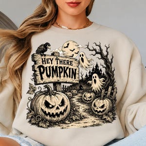 May include: A beige crewneck sweatshirt featuring a graphic print of a wooden sign that reads "HEY THERE PUMPKIN" with a black crow, a full moon, two ghosts, a haunted house, and three jack-o'-lanterns with carved faces in a dark forest setting.