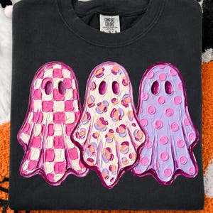 May include: A black crewneck sweatshirt featuring a graphic of three ghosts with textured, impasto-style paint strokes. The left ghost has a pink and white checkerboard pattern, the middle has a multi-colored leopard print, and the right is purple with pink polka dots.