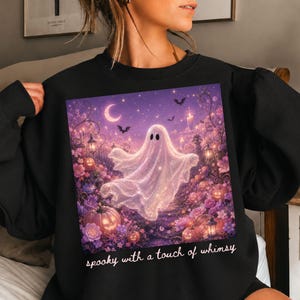 May include: A black sweatshirt featuring a graphic print of a glowing white ghost floating above a garden of purple flowers and orange pumpkins under a crescent moon and starry sky, with the text "spooky with a touch of whimsy" printed below.