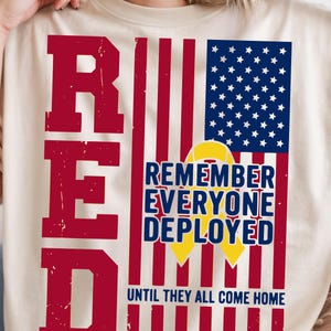 May include: Cream-colored t-shirt with a patriotic design. The design features the word "RED" in large, distressed red letters, alongside a stylized American flag and the text "REMEMBER EVERYONE DEPLOYED" with a yellow ribbon, and "UNTIL THEY ALL COME HOME."