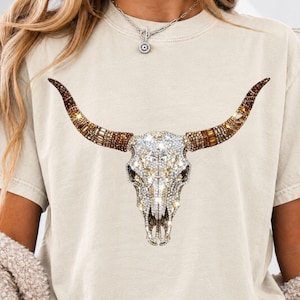 May include: A beige short-sleeve t-shirt featuring a large graphic of a bull skull. The skull is decorated with clear, sparkling rhinestones, while the long, curved horns are adorned with a mix of brown, amber, and gold-toned crystals.